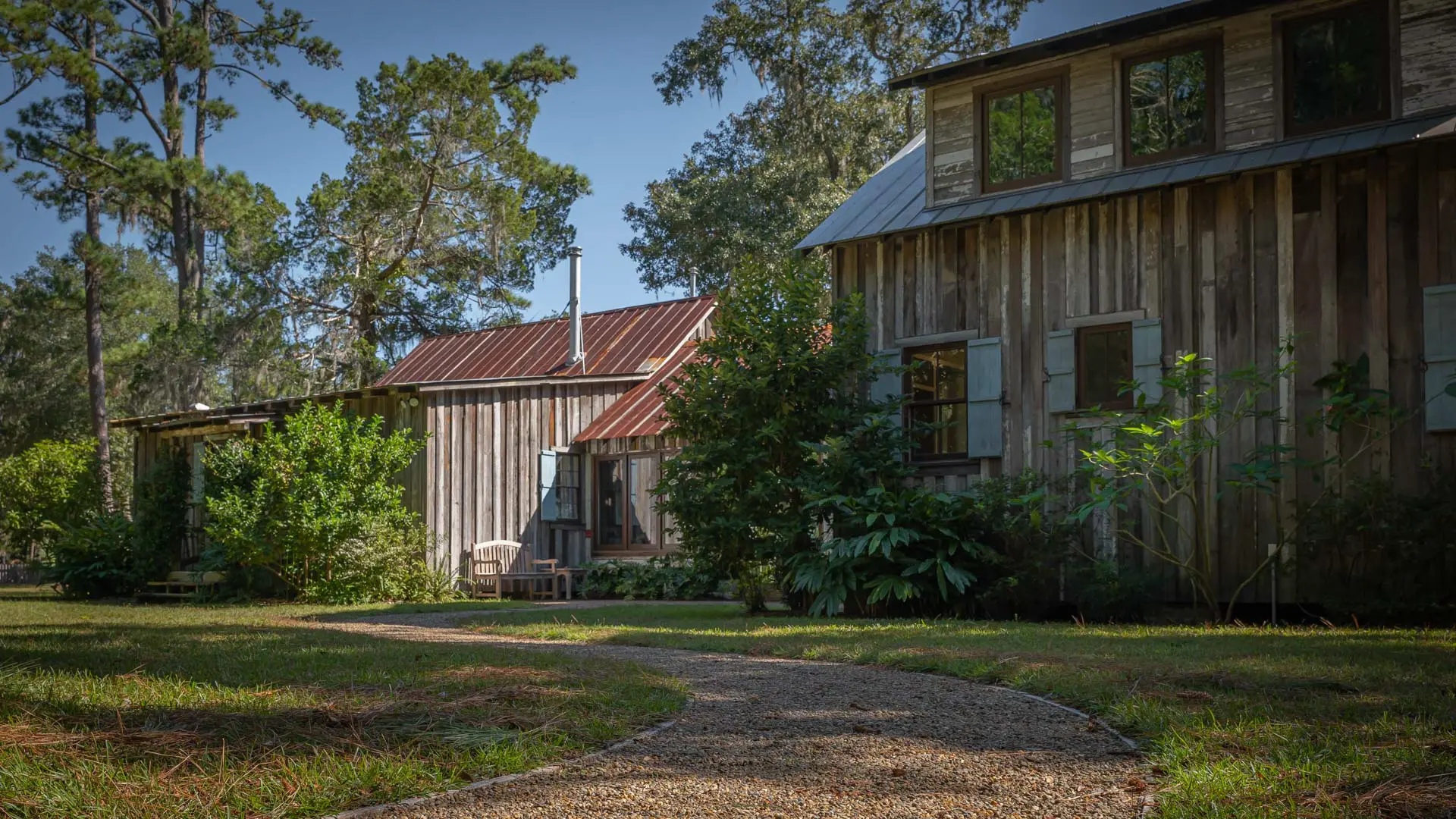 A gravel path curves through a grassy lawn toward two rustic, weathered wooden buildings surrounded by lush greenery and tall pine trees.