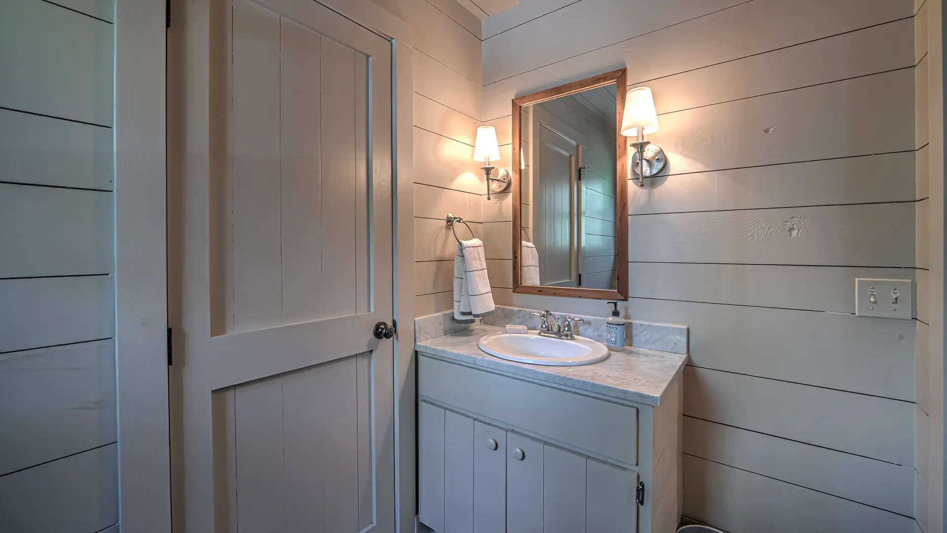 A white bathroom vanity with a white sink, a wood-framed mirror, and two wall sconces set against white shiplap walls.