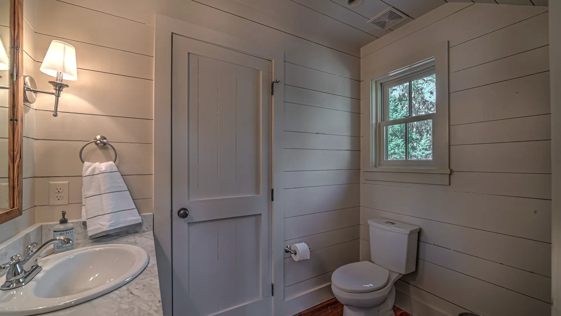 A bright powder room featuring white shiplap walls, a white sink with a marble countertop, and a white toilet positioned beneath a small window.