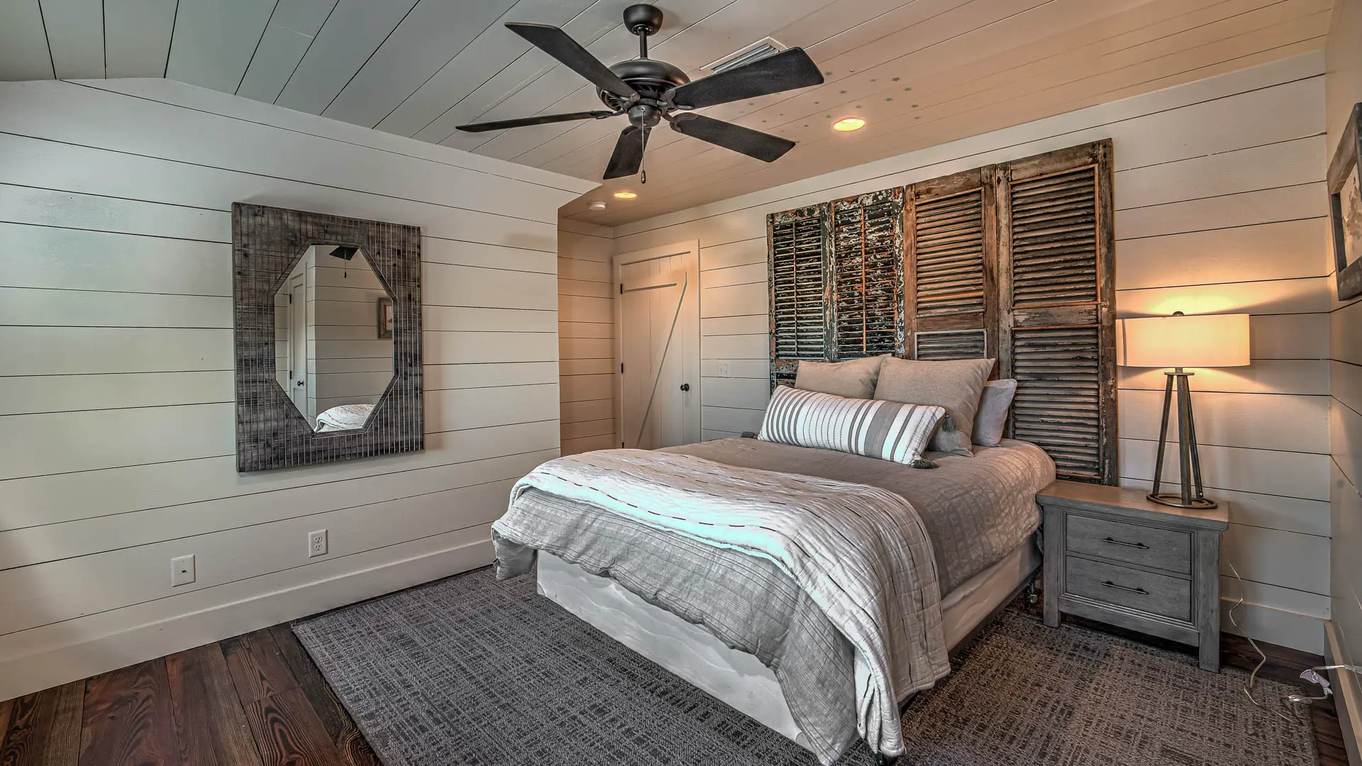 A rustic bedroom featuring white shiplap walls, a bed with a wooden shutter headboard, and a grey area rug on dark wood floors.