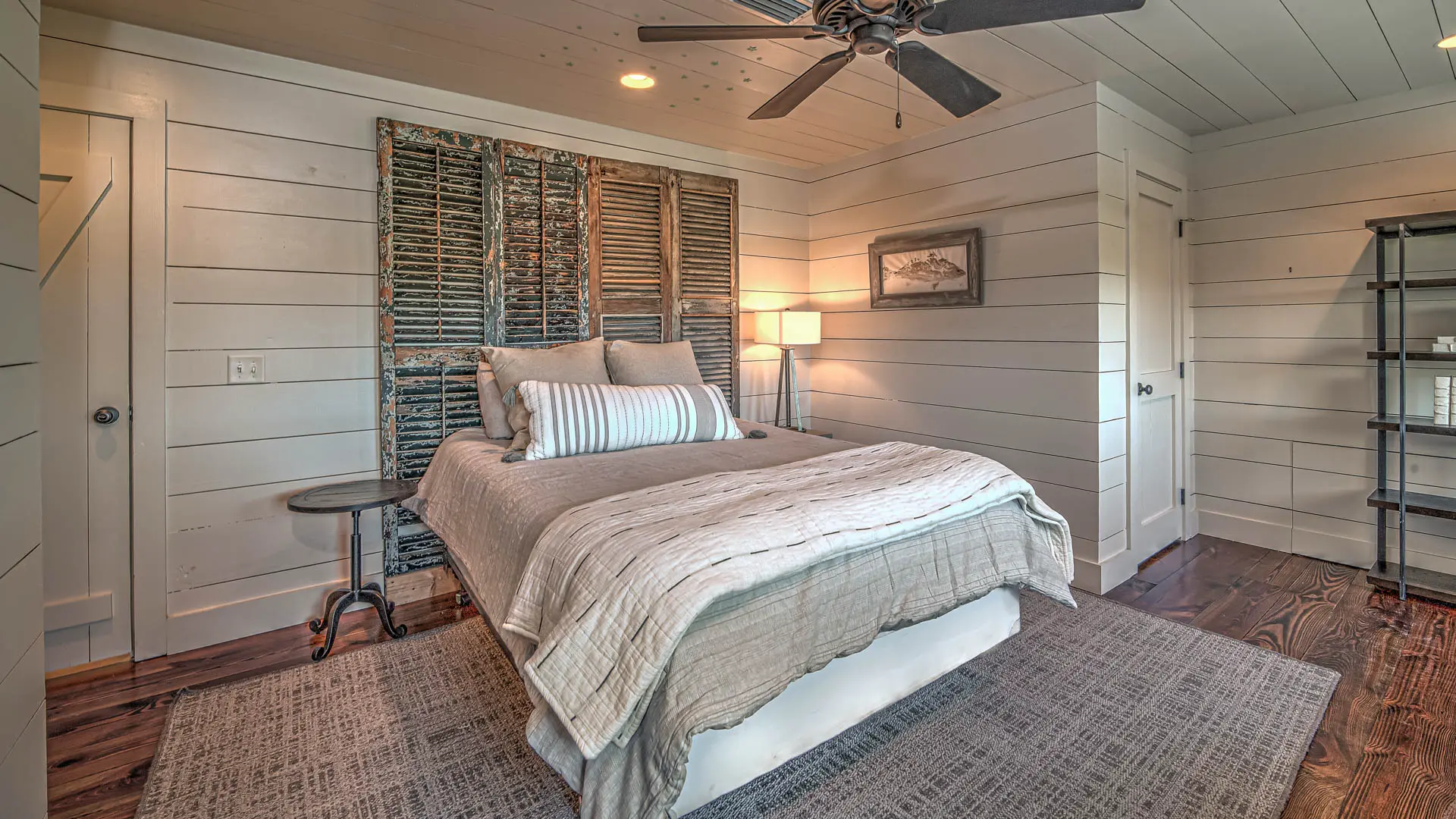 A cozy bedroom with white shiplap walls, a bed featuring a rustic reclaimed wood shutter headboard, and a grey area rug over dark hardwood floors.