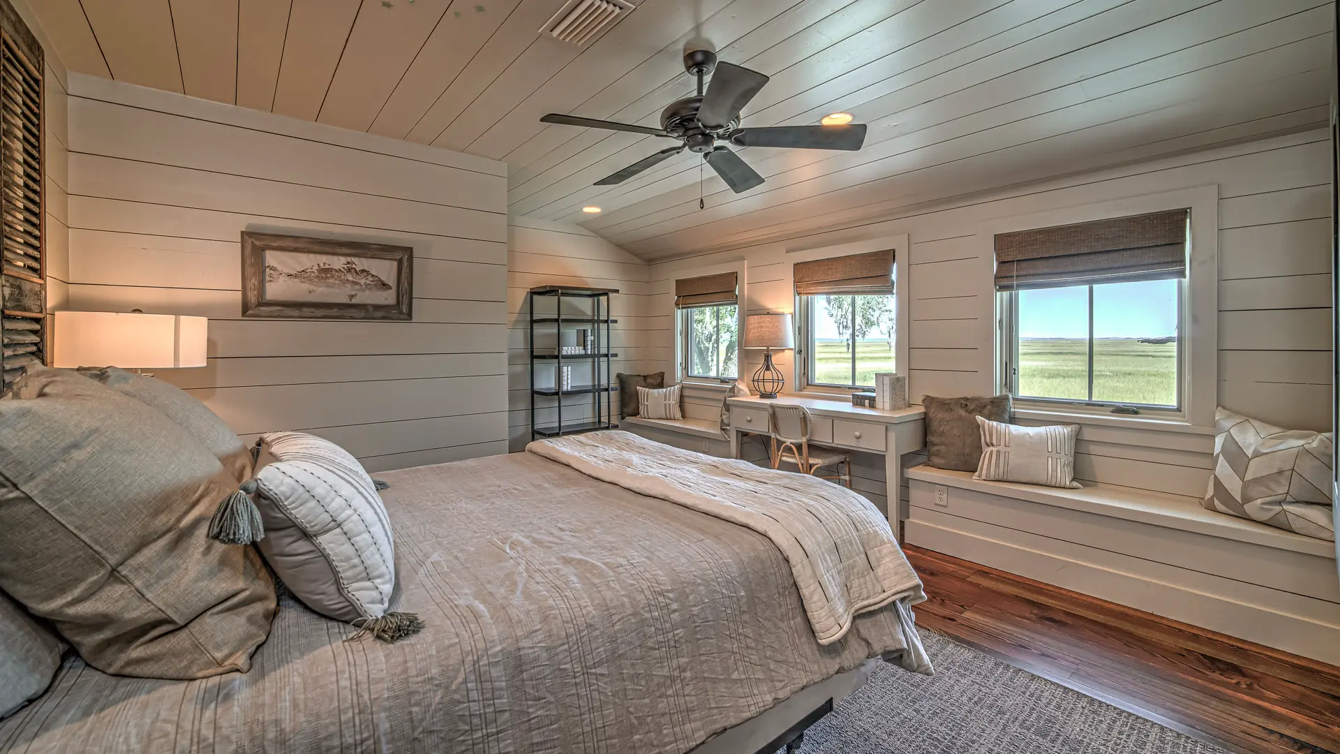 A serene bedroom with light-colored wood-paneled walls and ceiling, featuring a large bed with neutral linens, a built-in window seat, and a small desk.