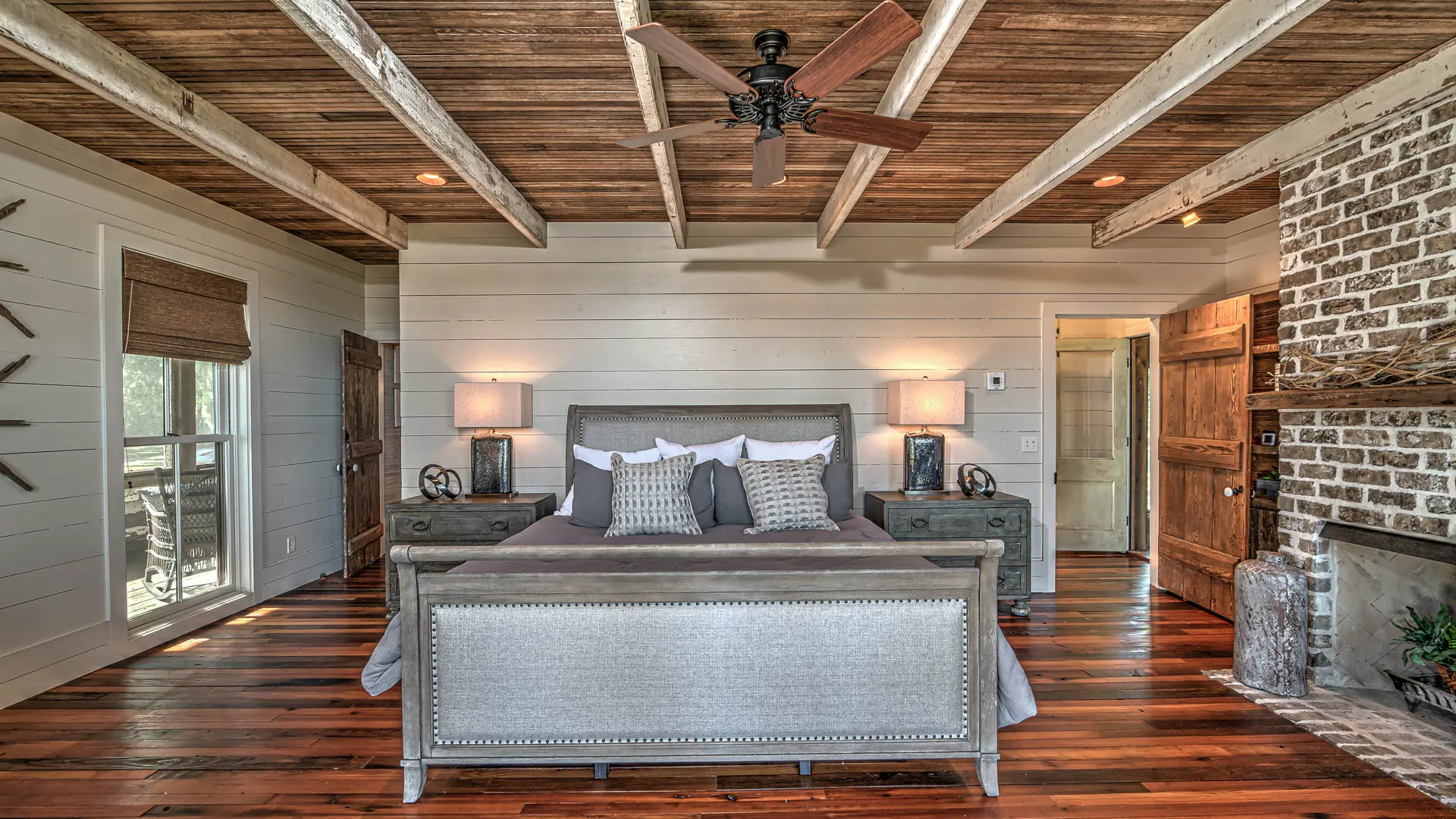 A rustic bedroom featuring white shiplap walls, polished hardwood floors, a wood-beamed ceiling, and a grey bed with a metallic footboard, flanked by nightstands and adjacent to a brick fireplace.