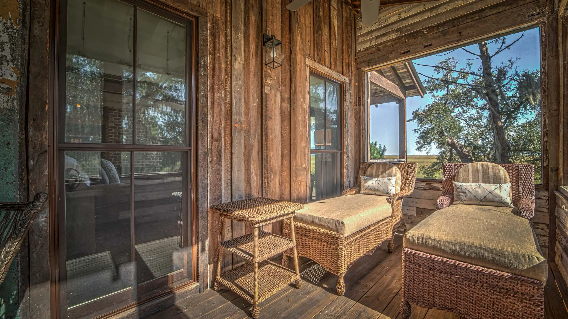 Two wicker lounge chairs with cushions and a small side table sit on a rustic wooden porch overlooking a sunny landscape.