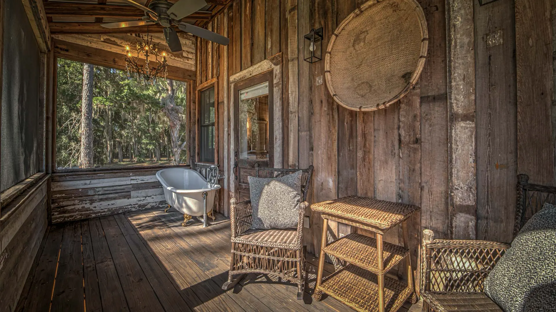 A rustic wooden room features a white clawfoot bathtub overlooking a lush forest, accompanied by wicker chairs and a small side table.