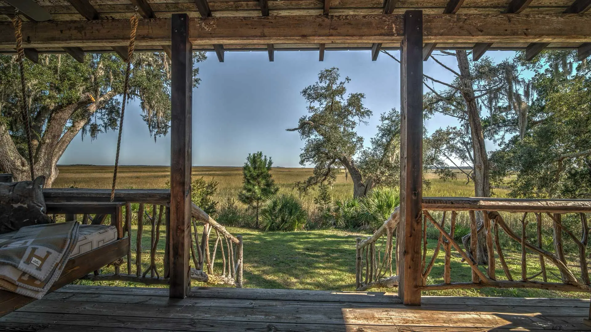 A rustic wooden porch with a swing framing a scenic view of a green lawn, oak trees, and a wide golden field under a clear sky.