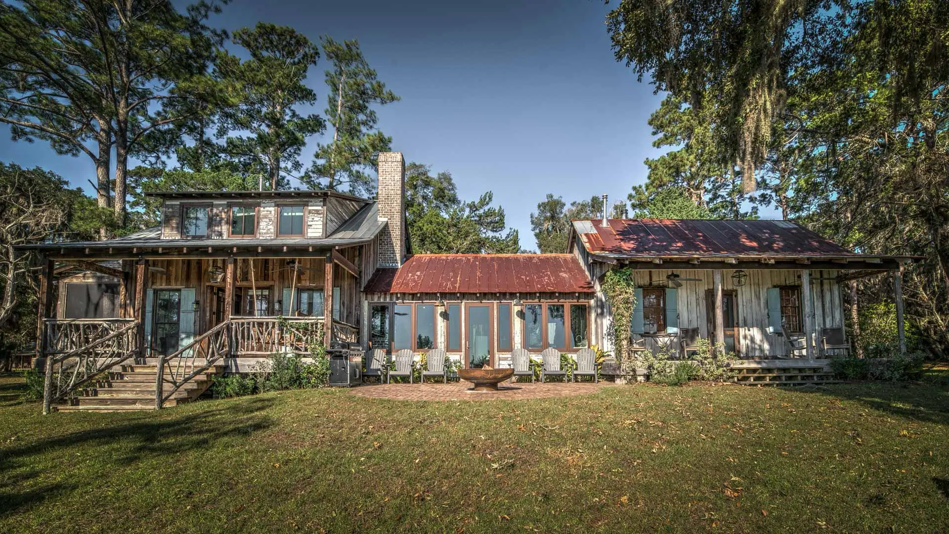 A sprawling rustic home with weathered red metal roofs and a wooden porch, set in a grassy yard surrounded by tall trees.