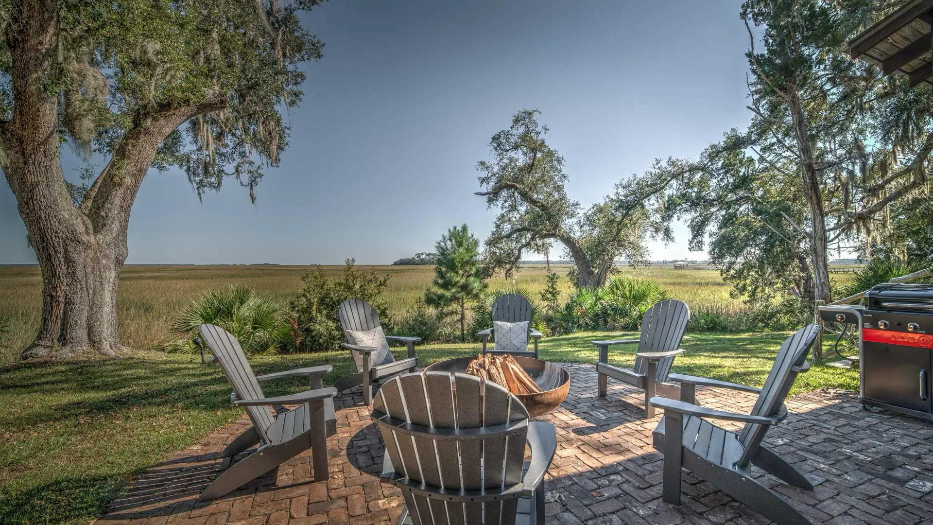 A cozy outdoor seating area with grey Adirondack chairs arranged around a fire pit on a brick patio, overlooking a wide marsh landscape.