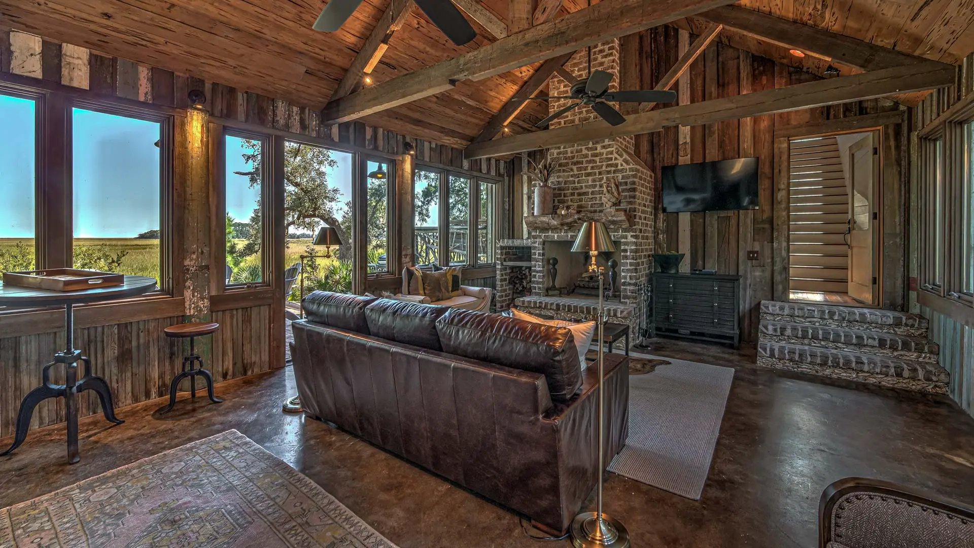 A rustic living room with wood-paneled walls and ceilings, featuring a large dark leather sofa, a brick fireplace, and large windows overlooking a scenic landscape.