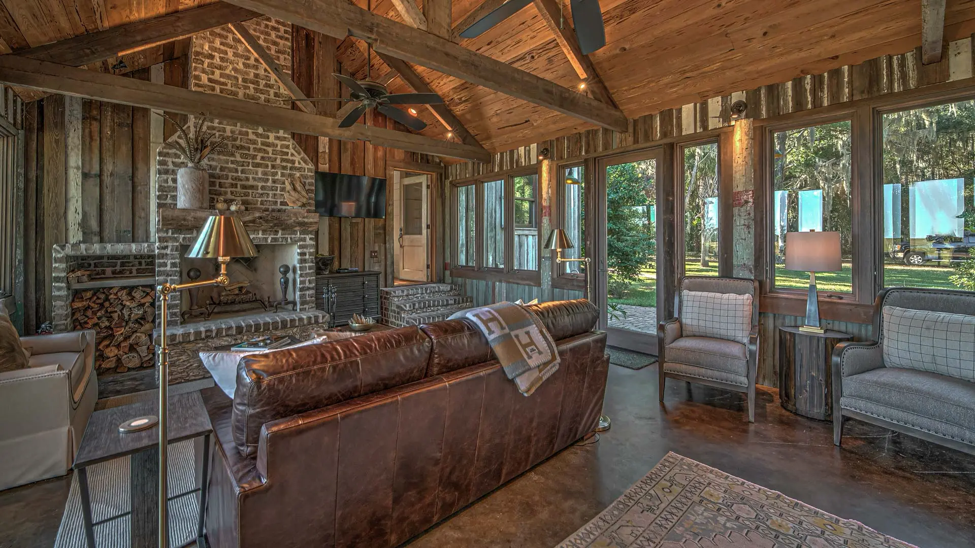 A cozy, rustic living room with high vaulted wooden beams, a brown leather sofa, and a large brick fireplace beside expansive windows overlooking a green landscape.