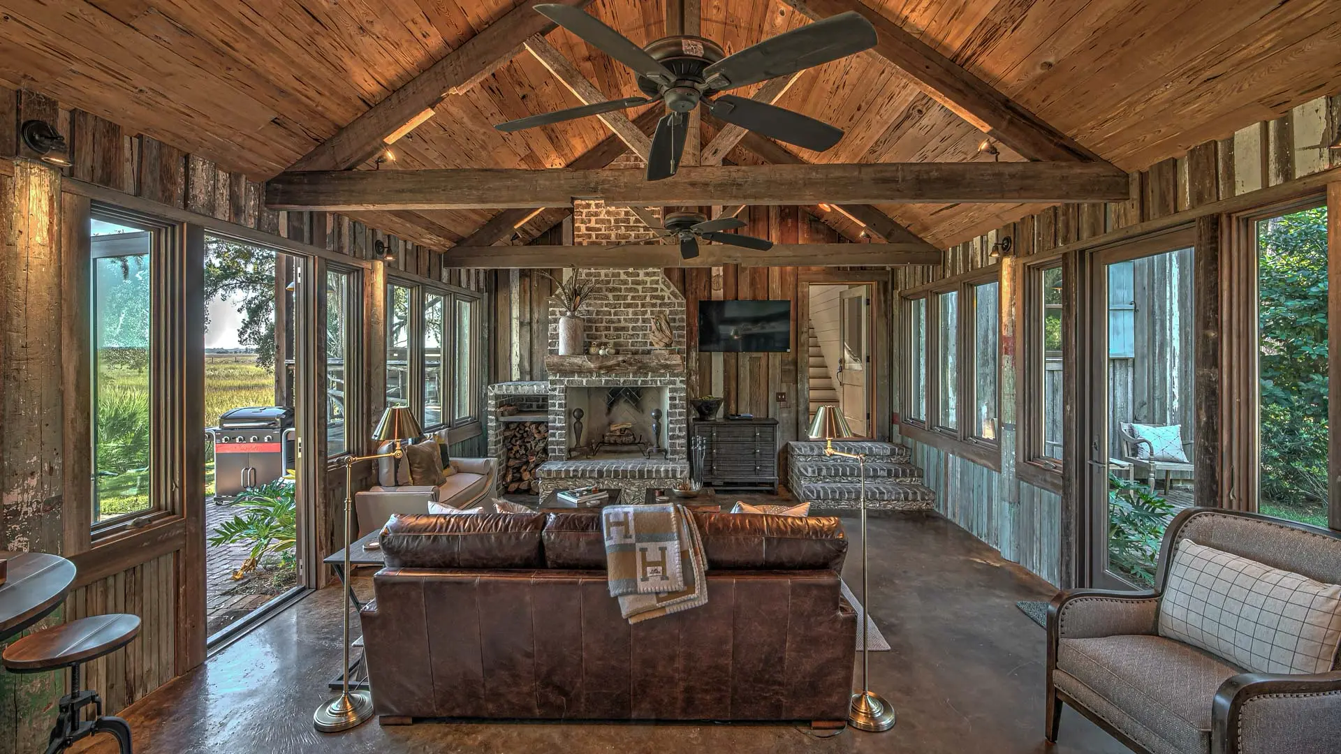 A cozy, rustic living room with vaulted wooden ceilings, a brown leather sofa, and a stone fireplace, surrounded by large windows that overlook a green landscape.