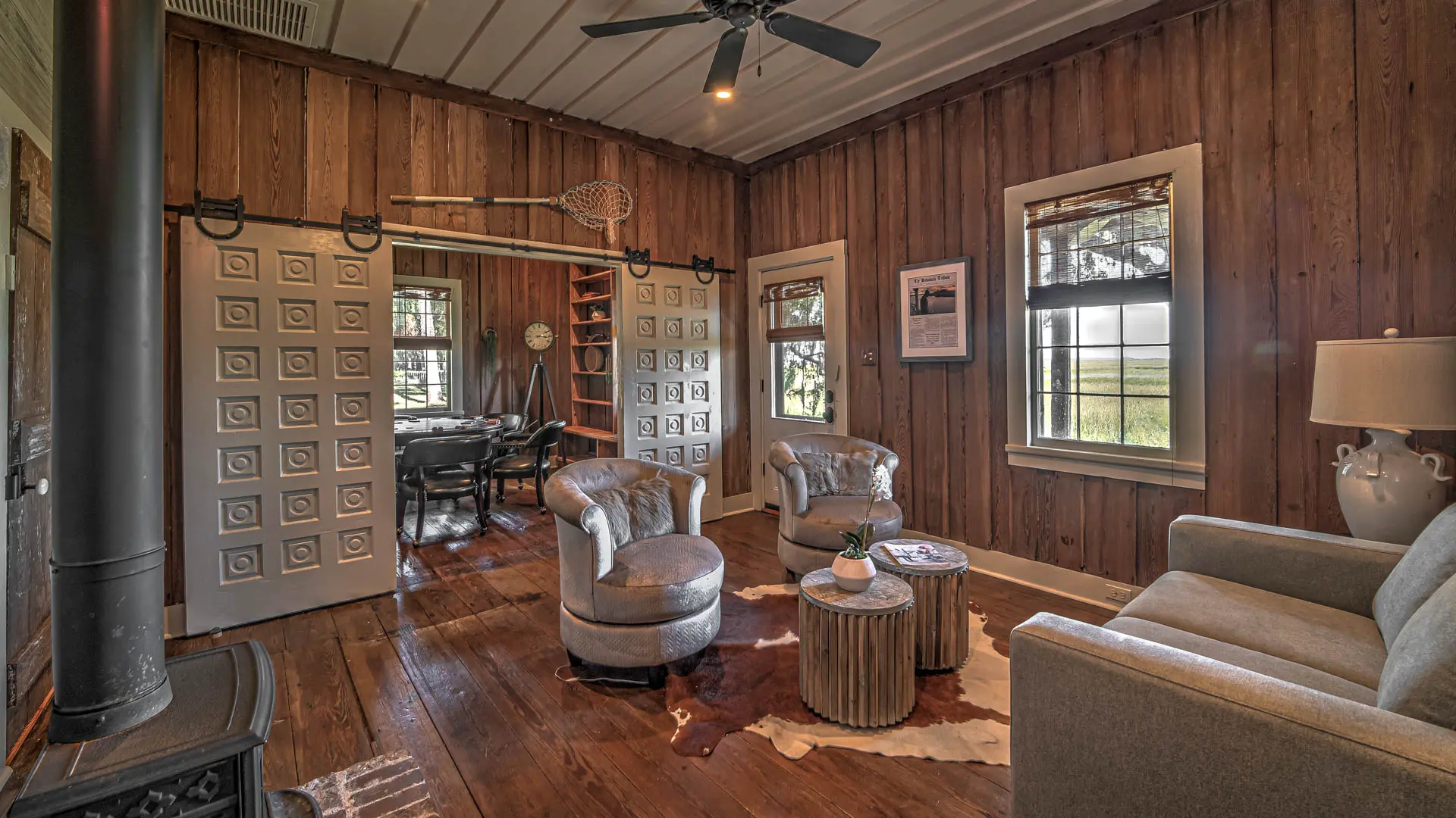 A rustic living room with wood-paneled walls and floors, featuring light-colored furniture, a black wood-burning stove, and white sliding barn doors that open into an adjacent room.