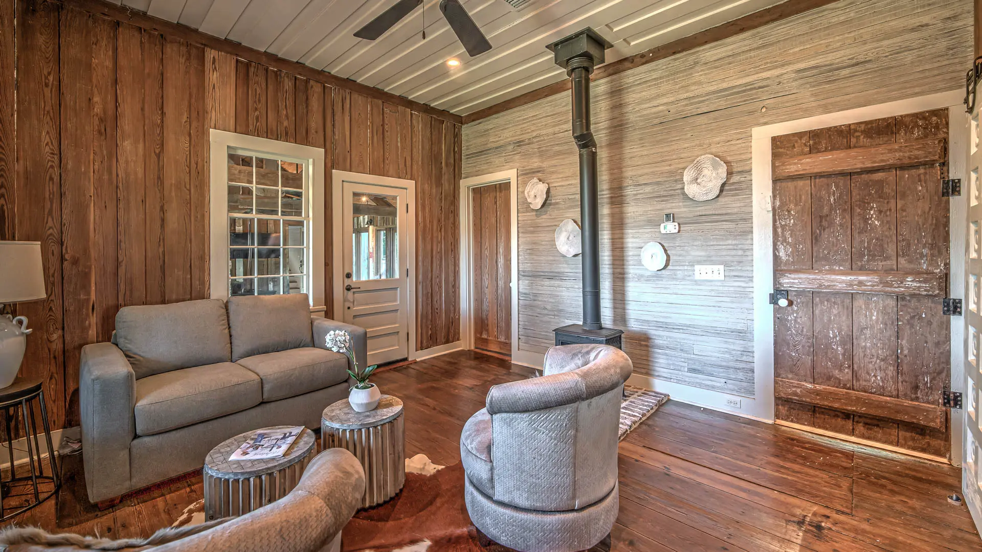 A rustic living room featuring wood-paneled walls, a gray sofa and armchair, and a black wood-burning stove.