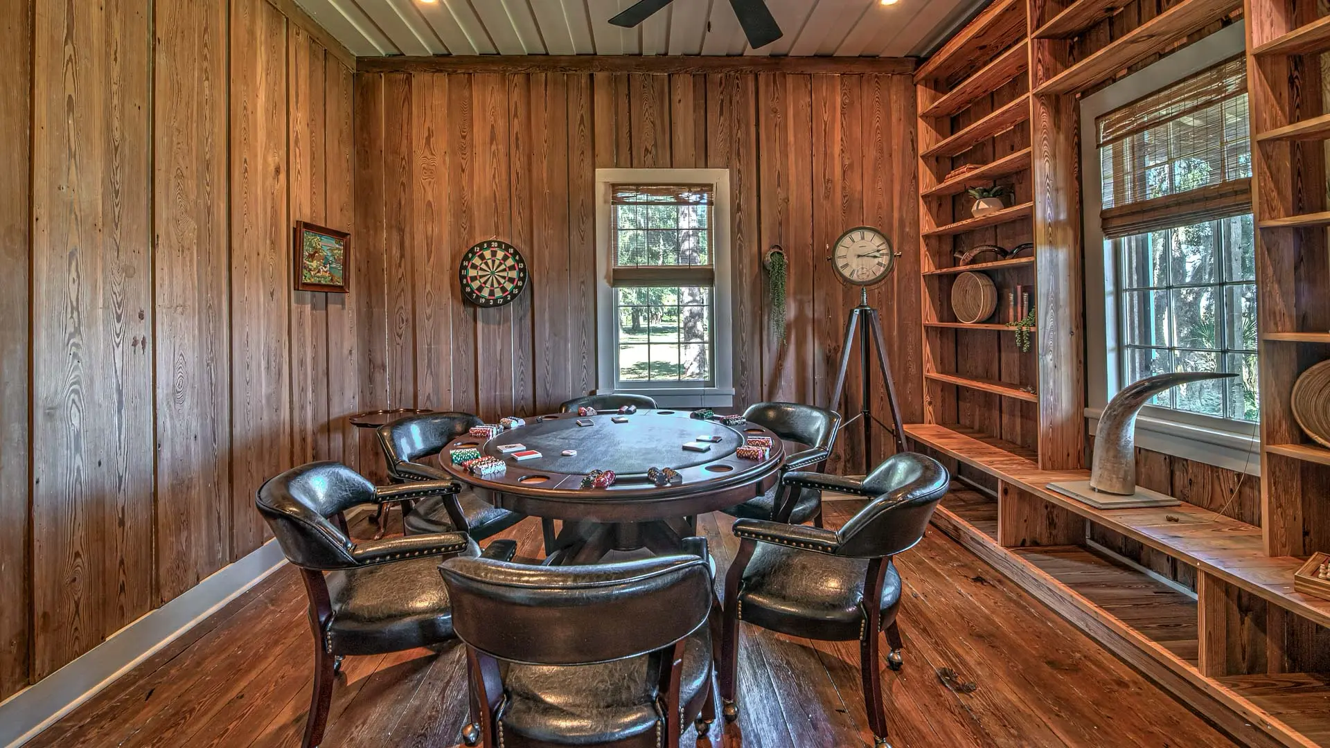 A rustic, wood-paneled game room featuring a round poker table with chips surrounded by black leather chairs, with a dartboard and built-in shelving in the background.