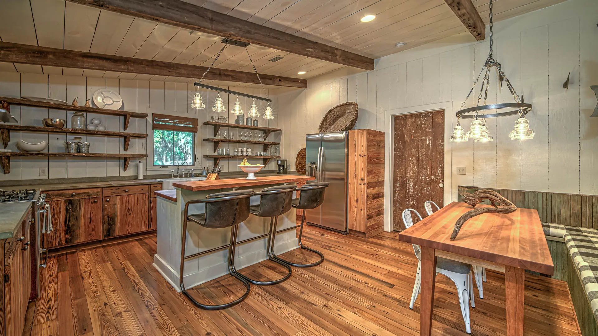 A rustic kitchen and dining space featuring wide-plank wooden floors, exposed ceiling beams, a center island with bar stools, and a wooden dining table.