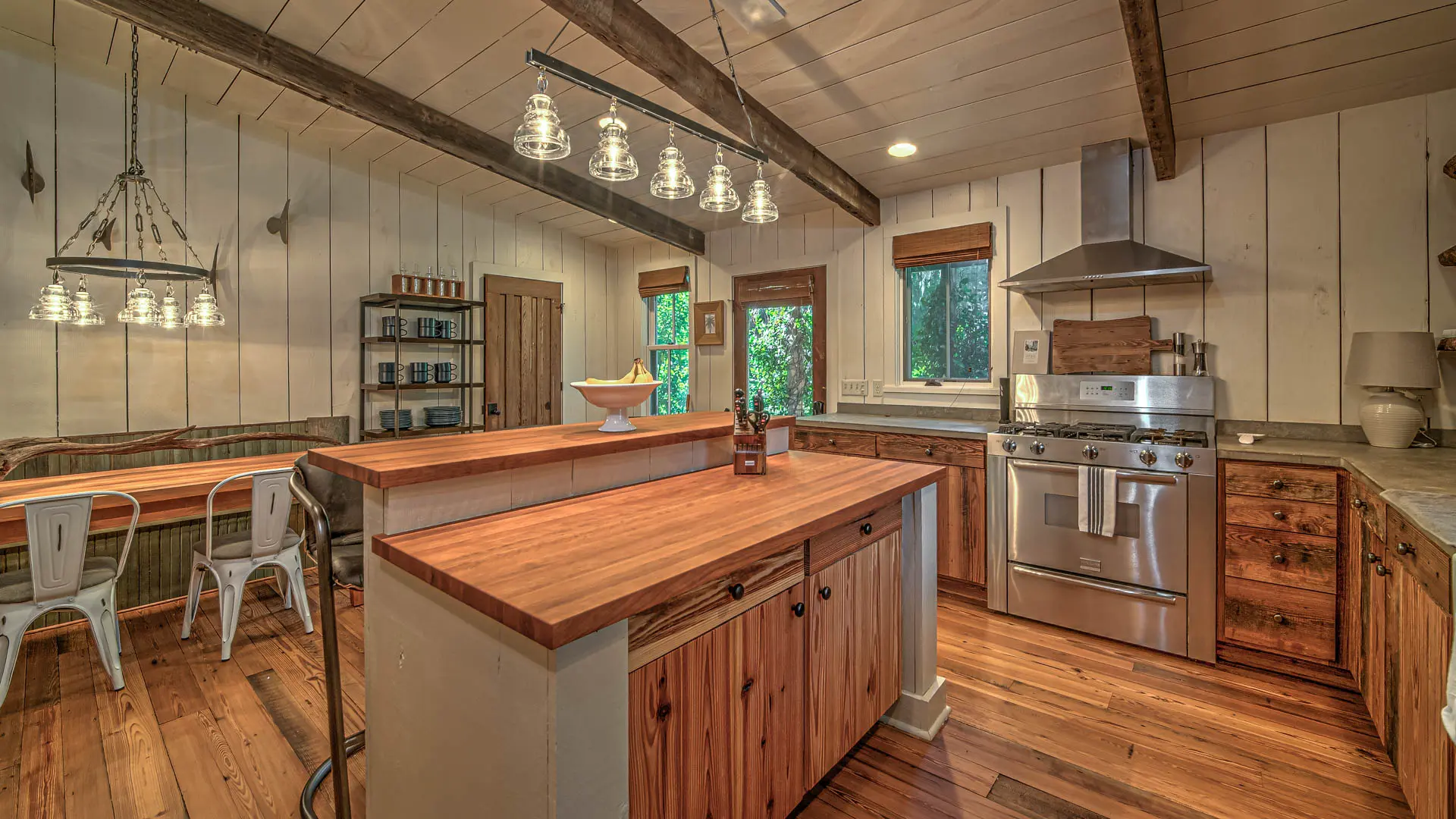 A rustic farmhouse kitchen featuring wooden countertops and flooring, white paneled walls, stainless steel appliances, and hanging light fixtures.