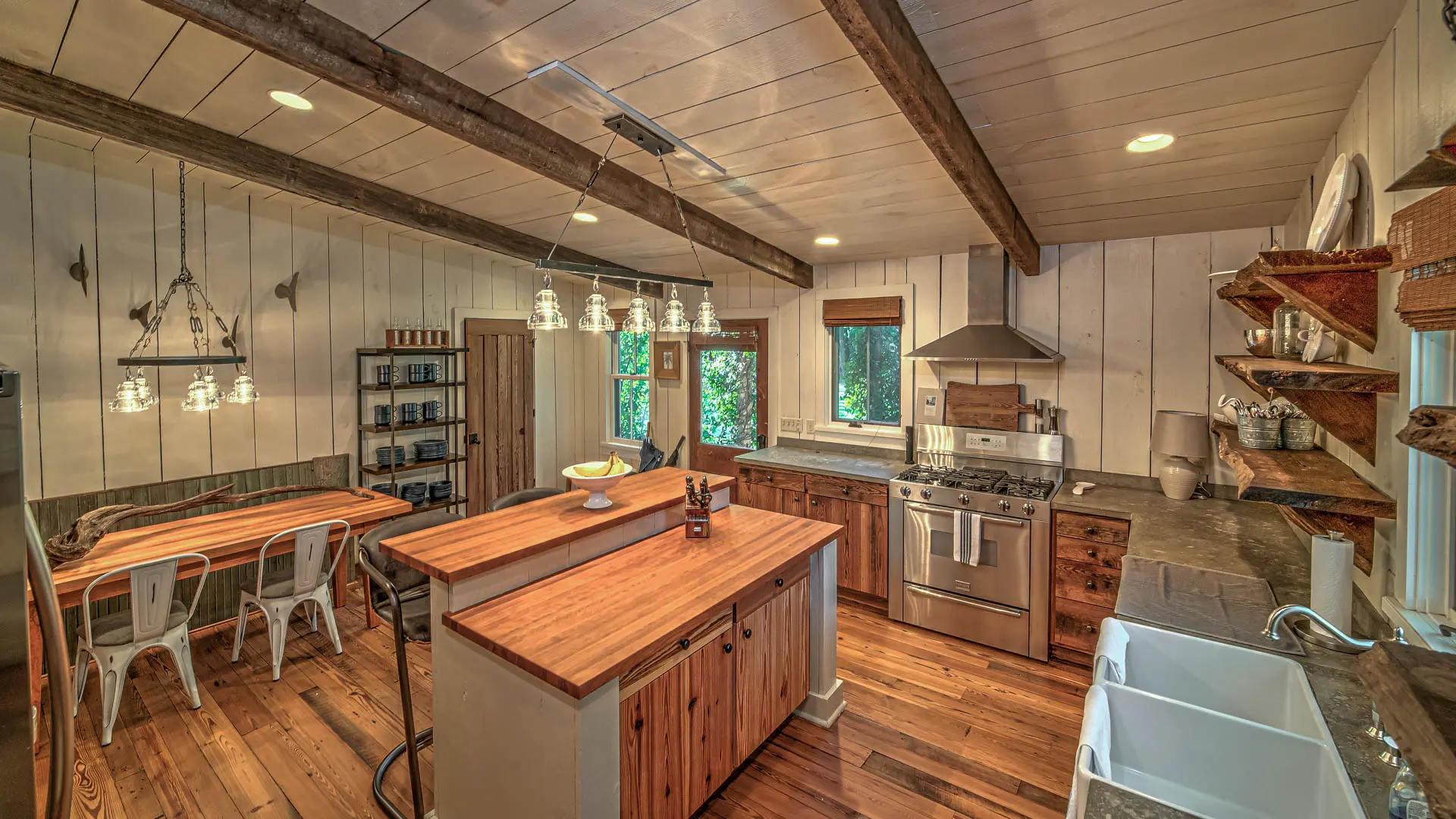 A cozy, rustic kitchen featuring white paneled walls, exposed wooden ceiling beams, wood countertops and flooring, and stainless steel appliances.