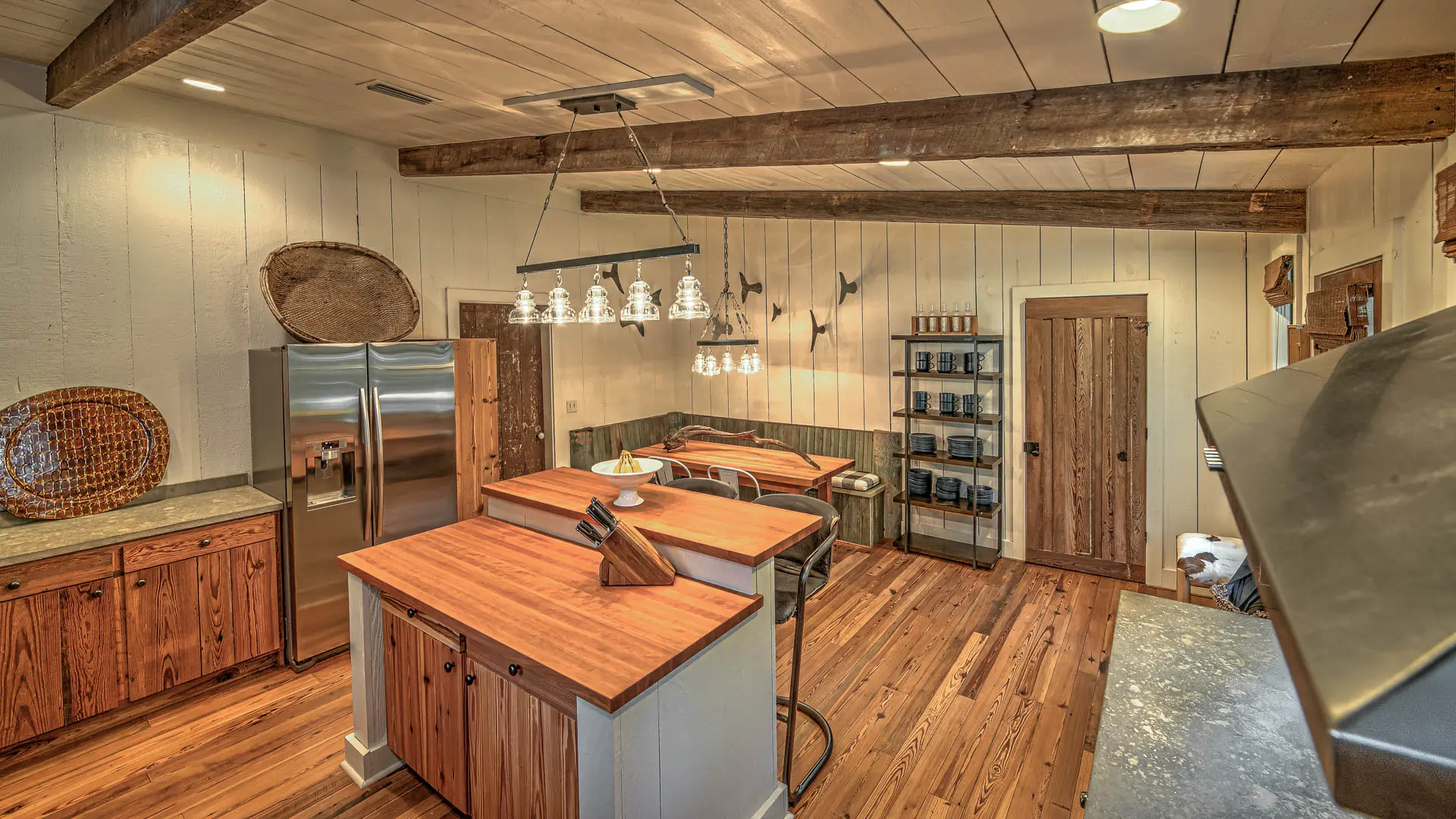 A rustic kitchen with light wood flooring, exposed ceiling beams, white paneled walls, a stainless steel refrigerator, and two wooden islands.