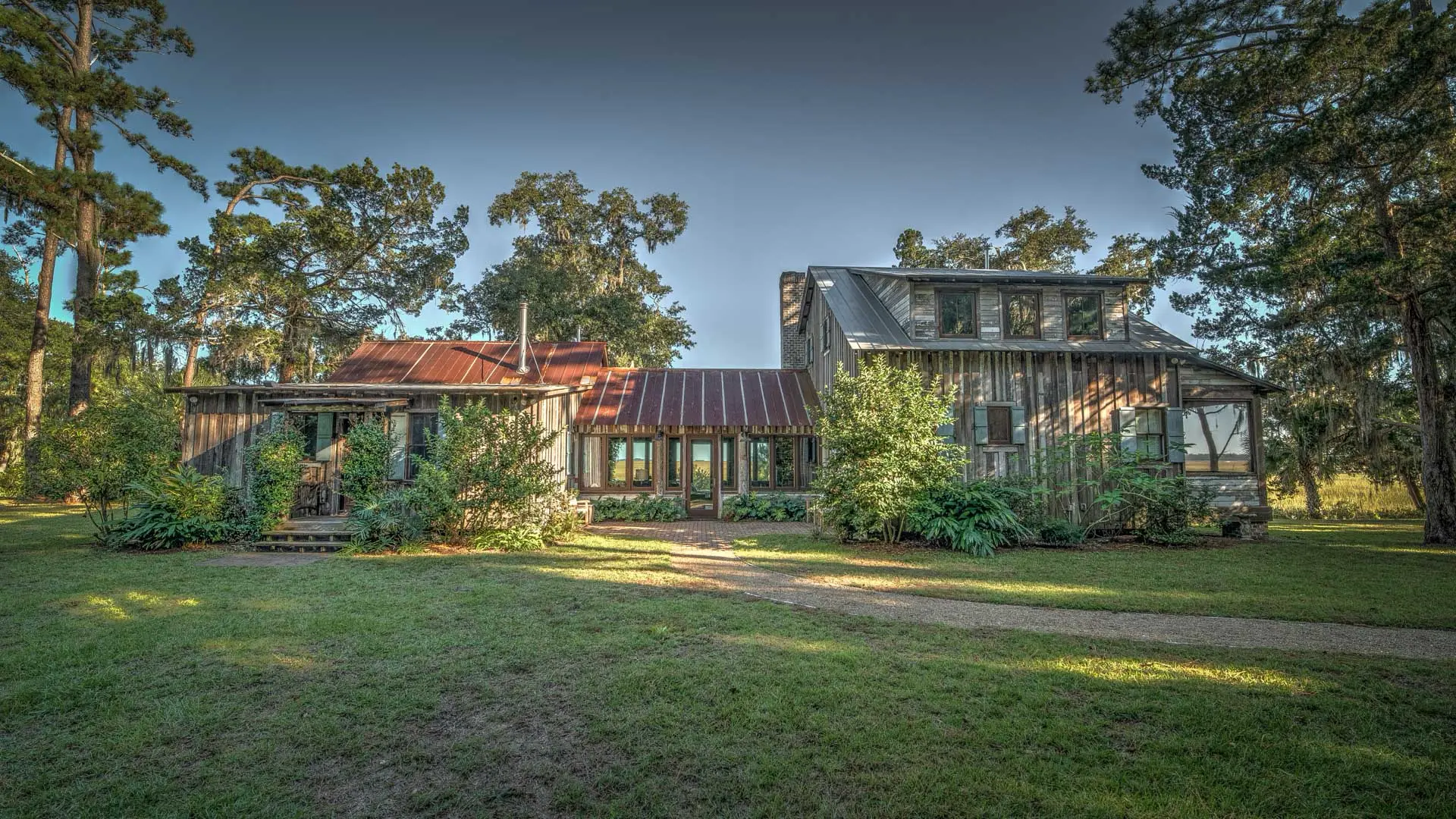 A rustic wooden house with corrugated rusted metal roofs, surrounded by lush green grass and tall trees.
