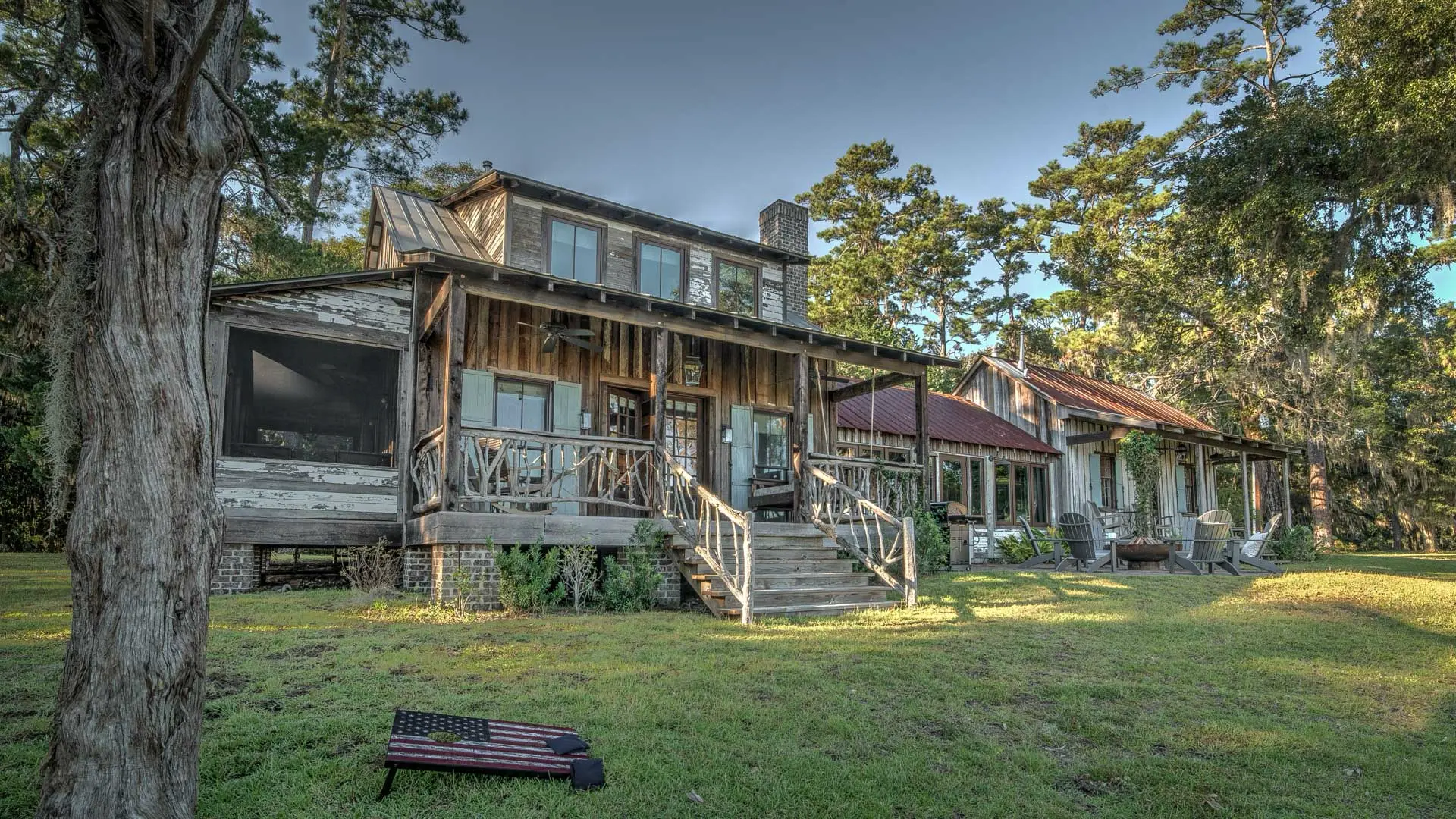 A rustic, weathered two-story wooden house with a front porch and wooden stairs, situated in a grassy clearing surrounded by tall trees.