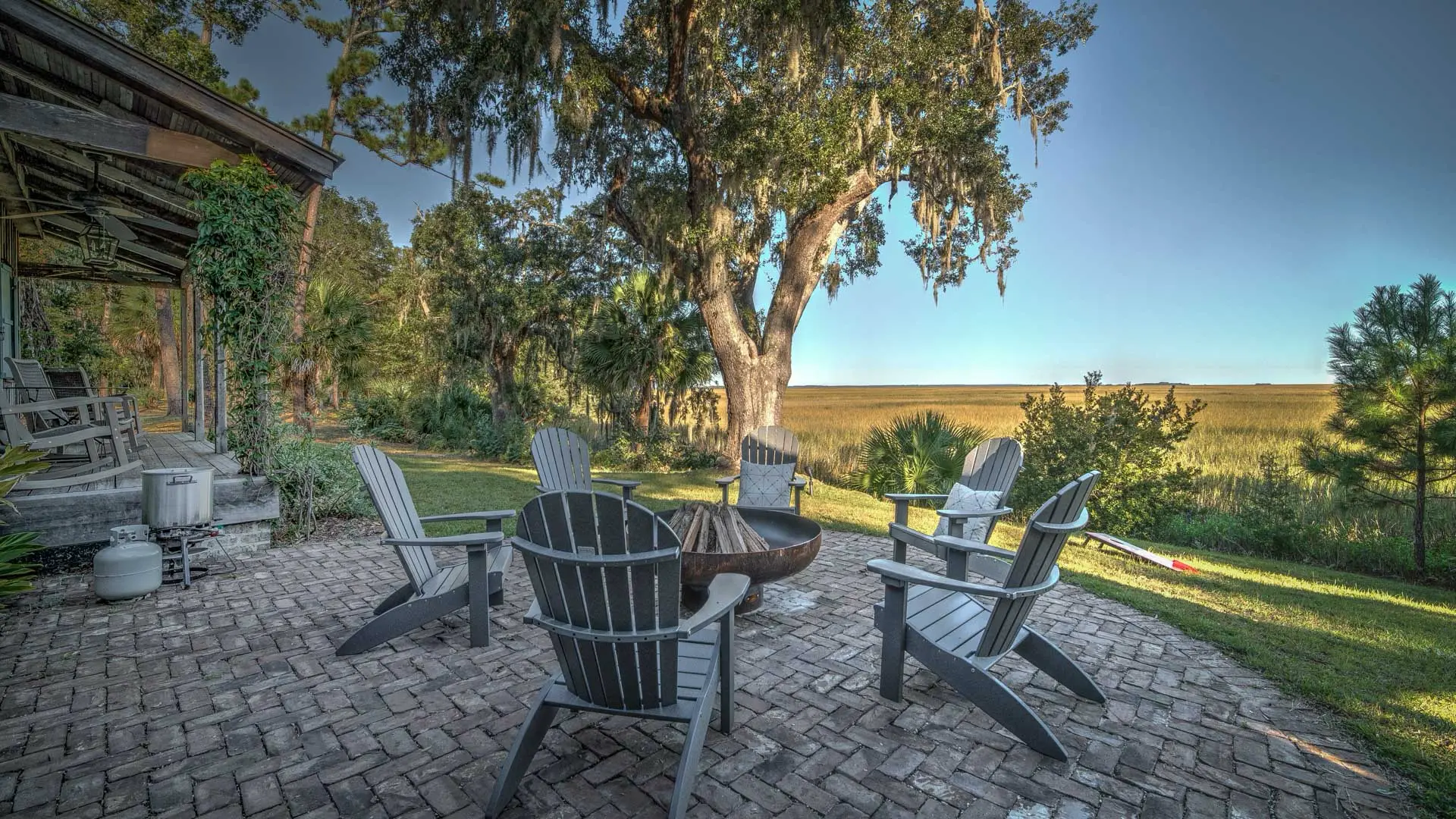 Gray Adirondack chairs surround a fire pit on a brick patio overlooking a scenic golden marsh under a clear blue sky.