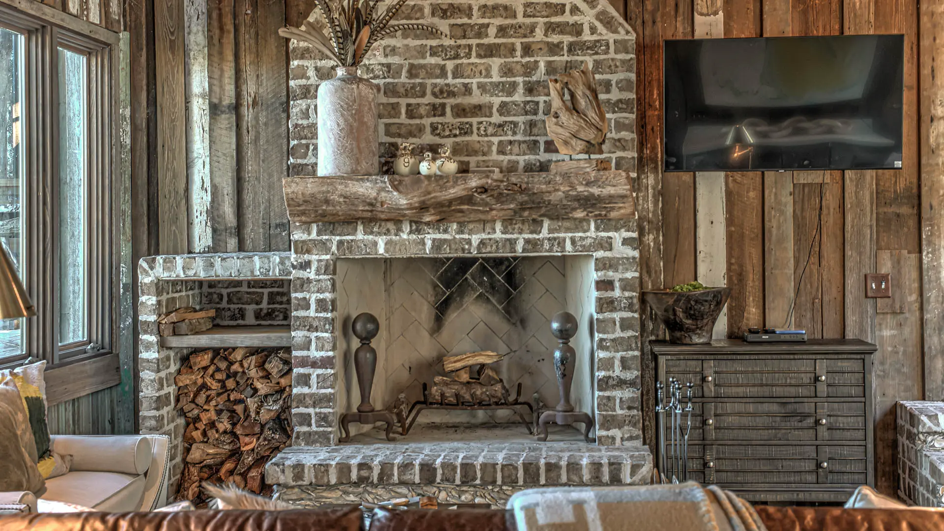 A rustic living room featuring a brick fireplace with a reclaimed wood mantel, flanked by a wood storage niche and a dark wooden dresser beneath a wall-mounted television.
