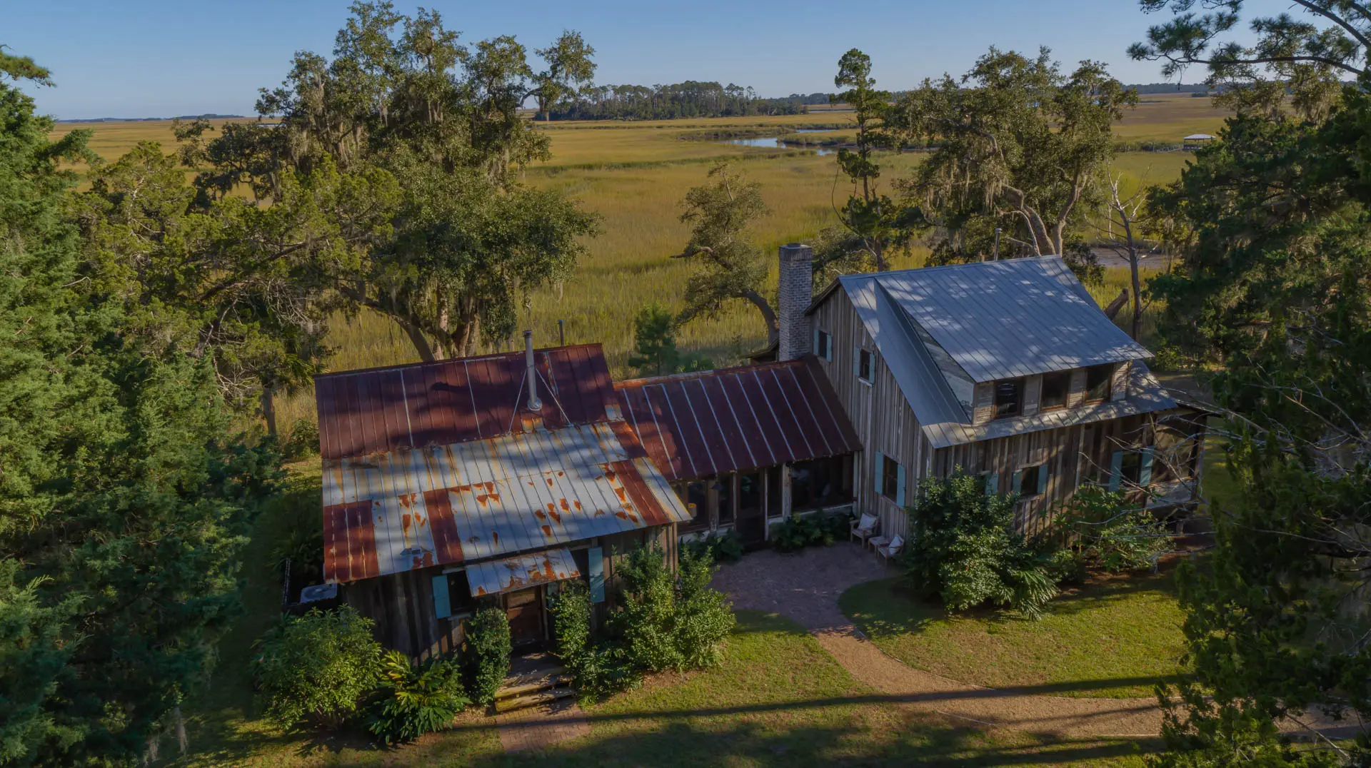 An aerial view of a rustic home with corrugated metal roofs, surrounded by lush green trees and overlooking a vast, open field.