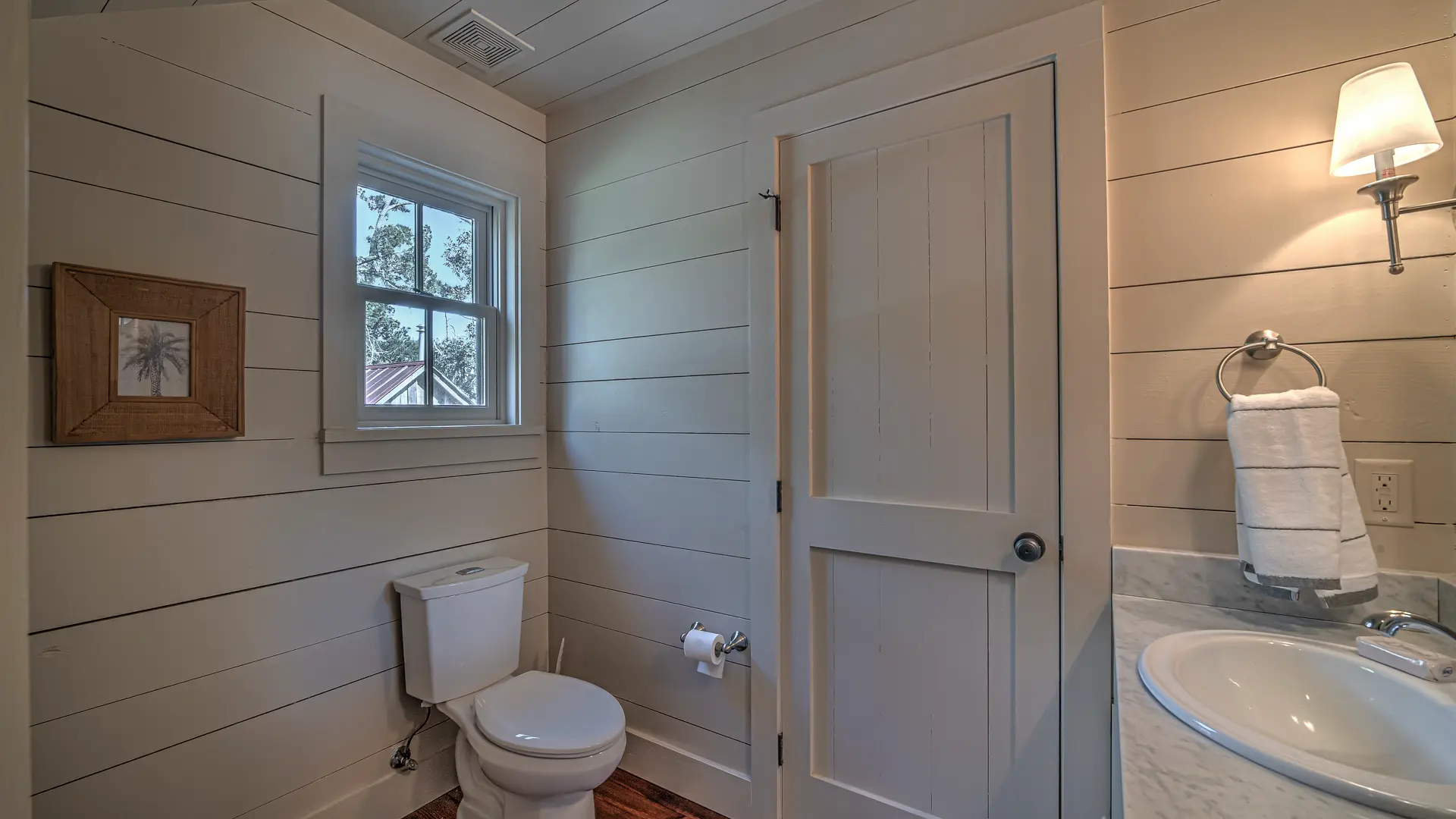 A small bathroom with light-colored shiplap walls, featuring a white toilet, a white sink vanity, and a small window.