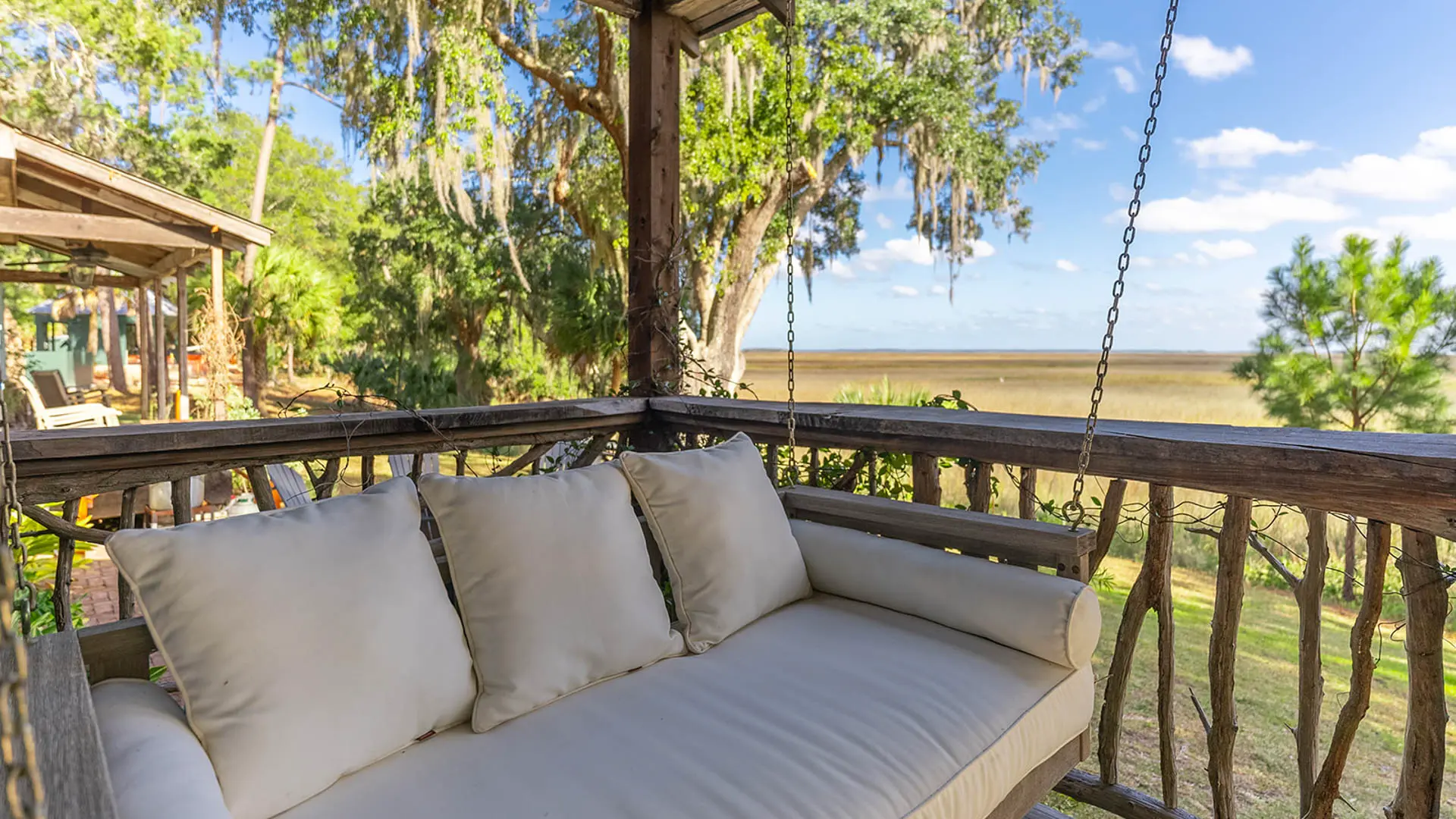 A white cushioned porch swing on a rustic wooden deck overlooking a serene marsh landscape with large trees draped in Spanish moss.