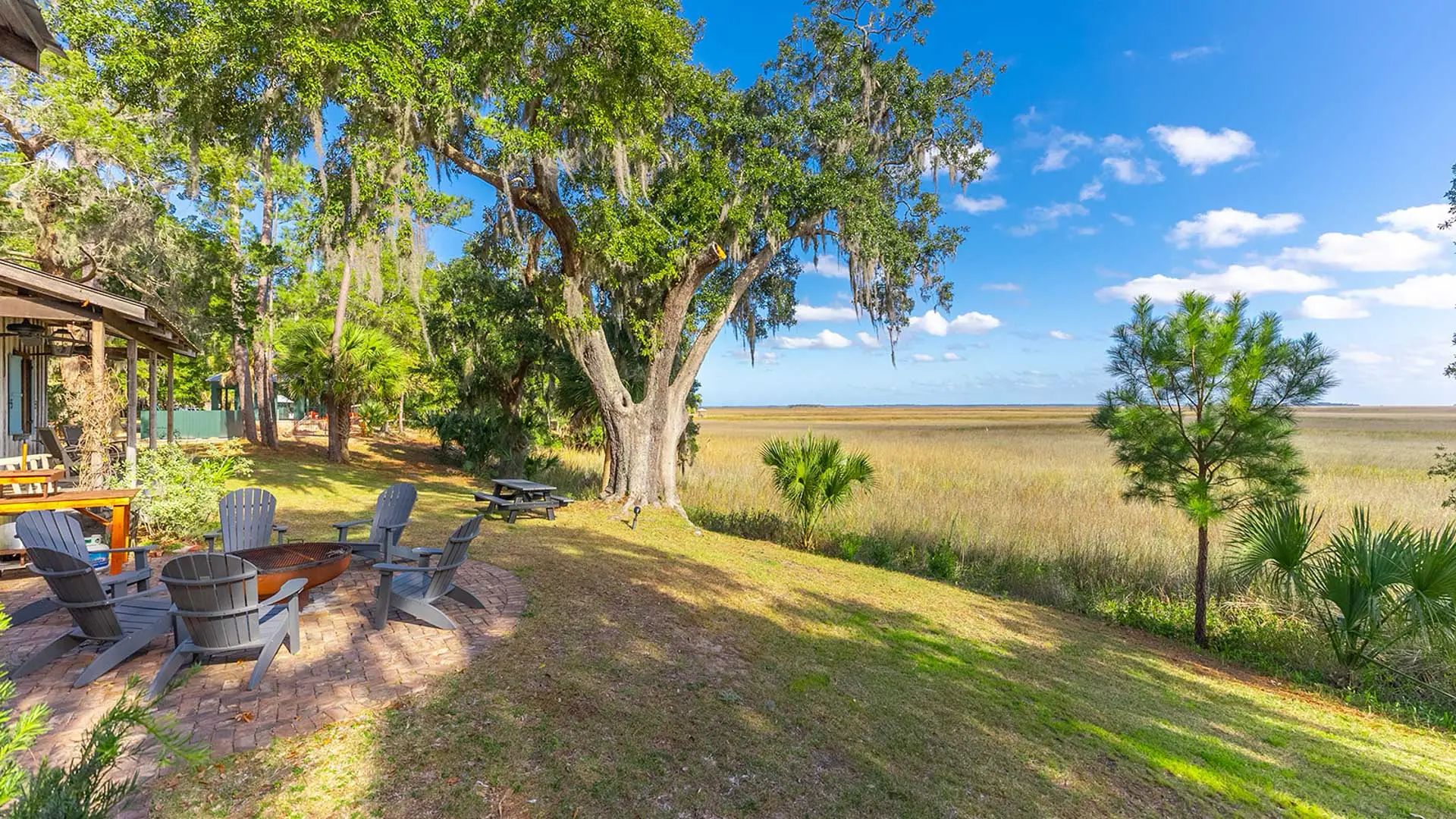 A sunny backyard featuring a fire pit area with grey Adirondack chairs, a large tree with Spanish moss, and a scenic view of a vast golden field under a bright blue sky.