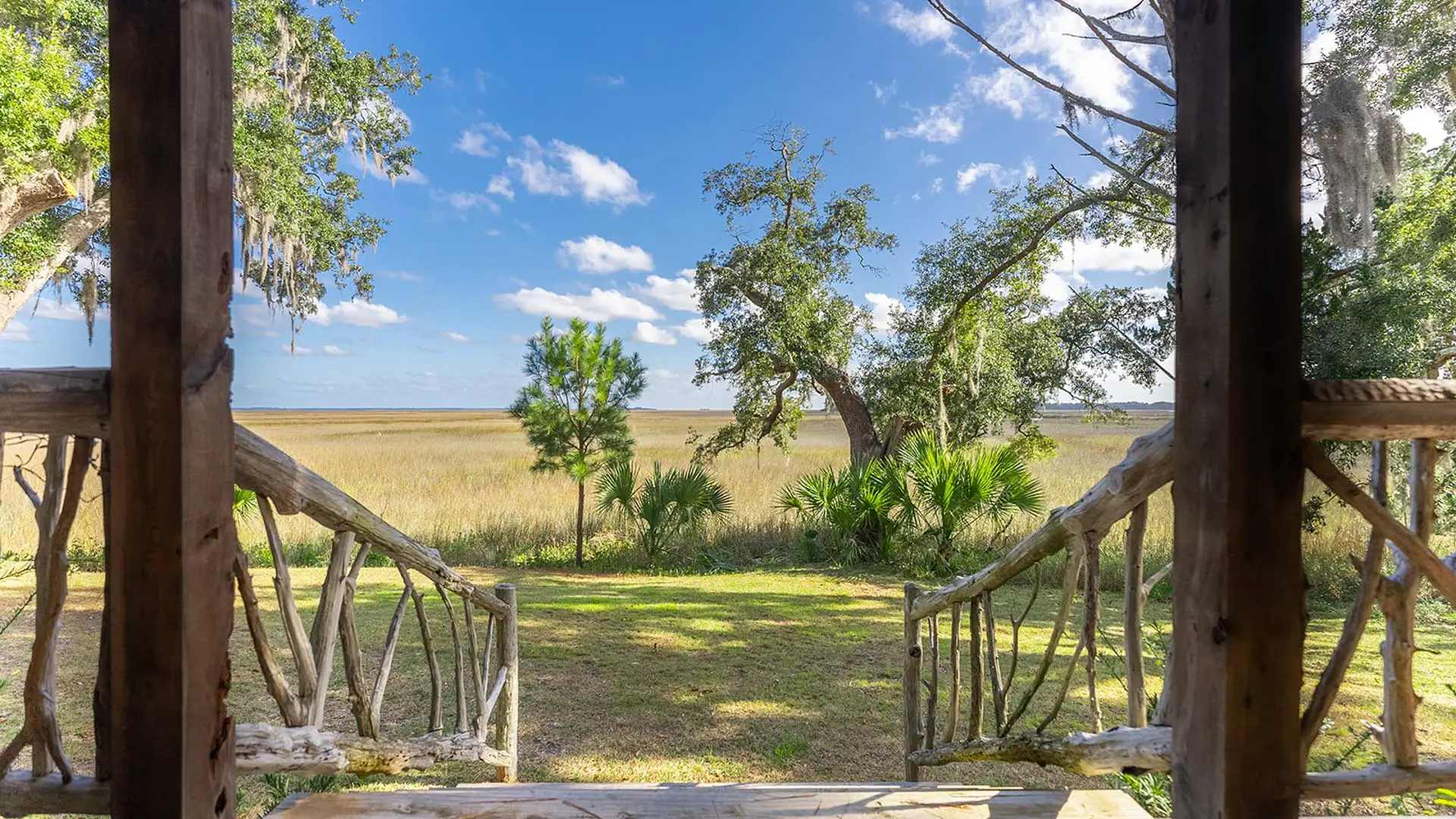 A view from a rustic wooden porch with branch railings looking out over a vast golden field and scattered trees under a bright blue sky with white clouds.