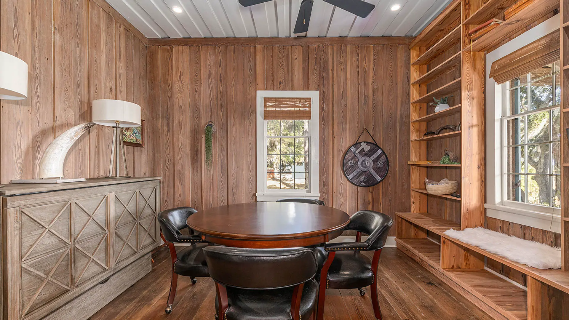 A rustic wood-paneled room featuring a round wooden table with black leather chairs, a textured grey sideboard, and built-in wooden shelving.