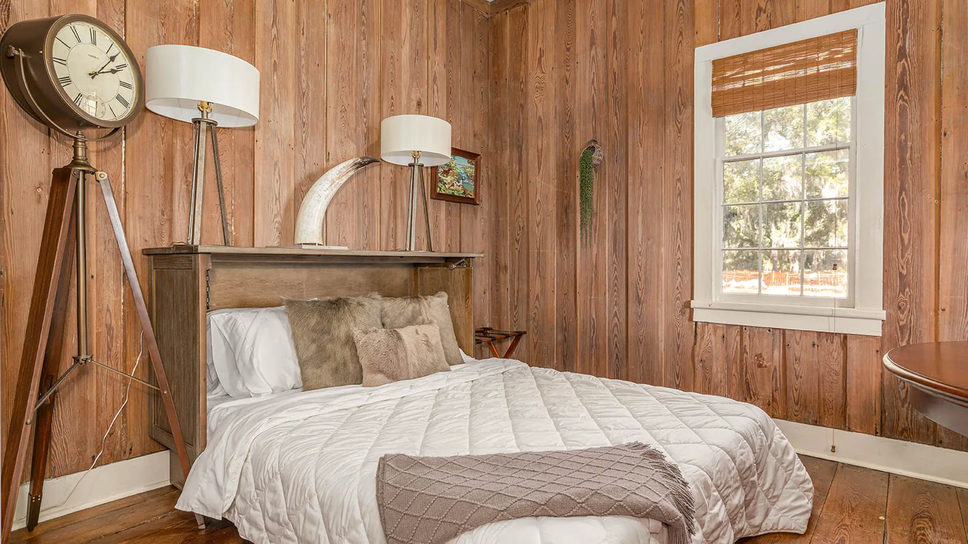 A cozy rustic bedroom with vertical wood-paneled walls, a bed with a white quilted comforter, and a large vintage-style clock on a wooden tripod stand.